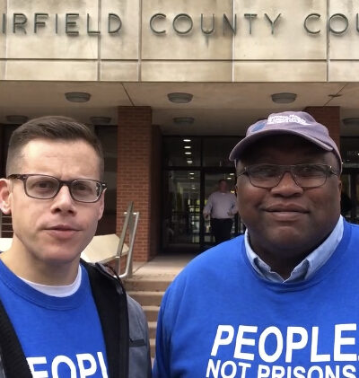ACLU-CT Gus Marks-Hamilton and Anderson Curtis in blue "people, not prisons" shirts, stand outside of the Fairfield County Courthouse