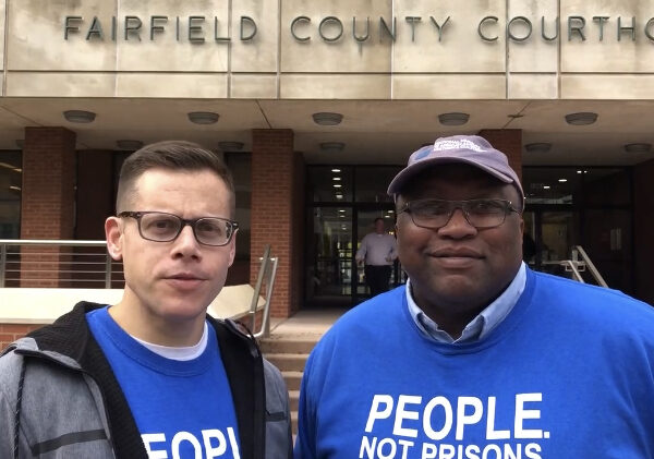 ACLU-CT Gus Marks-Hamilton and Anderson Curtis in blue "people, not prisons" shirts, stand outside of the Fairfield County Courthouse