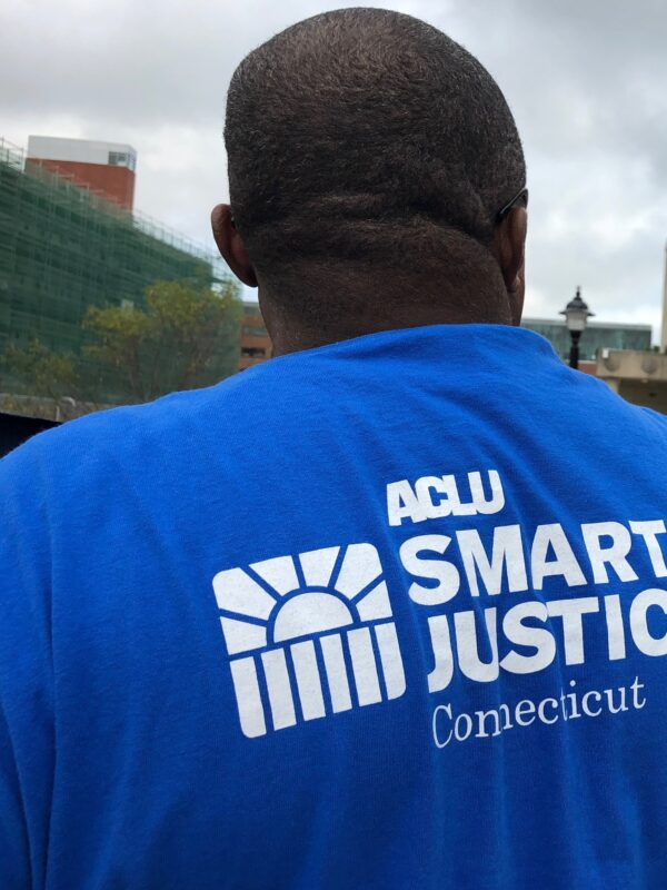 From behind, a man stands in a blue ACLU of Connecticut Smart Justice shirt