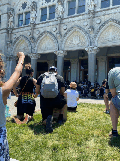 A crowd of people, spaced apart for physical distancing, take a knee in front of the Connecticut capitol building. A person in the foreground, hair up in a ponytail, holds her fist up.