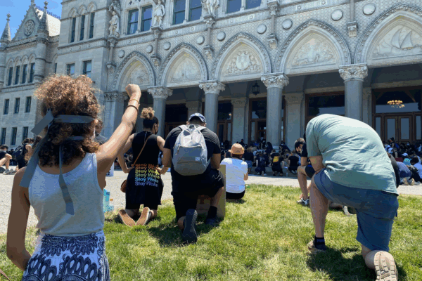 A crowd of people, spaced apart for physical distancing, take a knee in front of the Connecticut capitol building. A person in the foreground, hair up in a ponytail, holds her fist up.