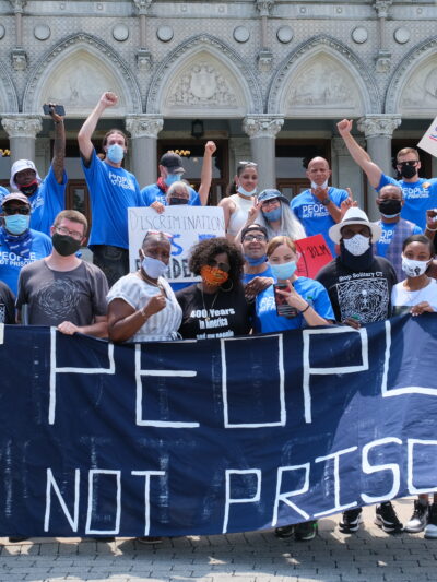 A group of Smart Justice leaders, advocates, and legislators, all wearing masks, stand behind the ACLU-CT's blue "People Not Prisons" banner outside of the Connecticut state capitol building.