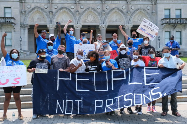 A group of Smart Justice leaders, advocates, and legislators, all wearing masks, stand behind the ACLU-CT's blue "People Not Prisons" banner outside of the Connecticut state capitol building.