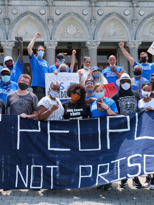 A group of Smart Justice leaders, advocates, and legislators, all wearing masks, stand behind the ACLU-CT's blue "People Not Prisons" banner outside of the Connecticut state capitol building.