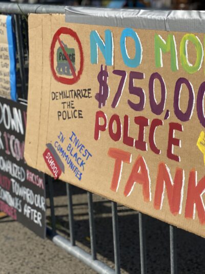 A cardboard sign taped to a fence outside of a Stamford Connecticut protest. The sign says: "No more $750,000 police tanks," "demilitarize the police," and "invest in our communities." It has a drawing of a raised fist, and of a tank crossed out.