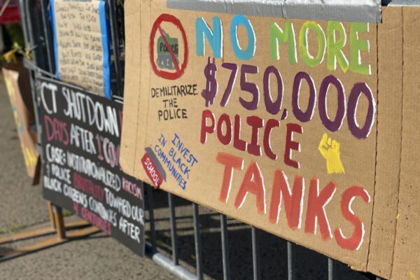 A cardboard sign taped to a fence outside of a Stamford Connecticut protest. The sign says: "No more $750,000 police tanks," "demilitarize the police," and "invest in our communities." It has a drawing of a raised fist, and of a tank crossed out.