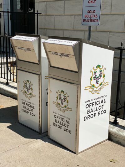 Two official Connecticut ballot drop boxes sit outside of the Waterbury city hall in 2020. The boxes are large, rectangular, and look like library book drops, with the state of CT seal visible, and "official ballot drop box," on their fronts and sides