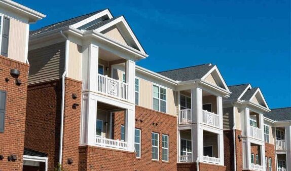 A block of apartments, white on top with balconies and brick below, stand in front of a blue sky.