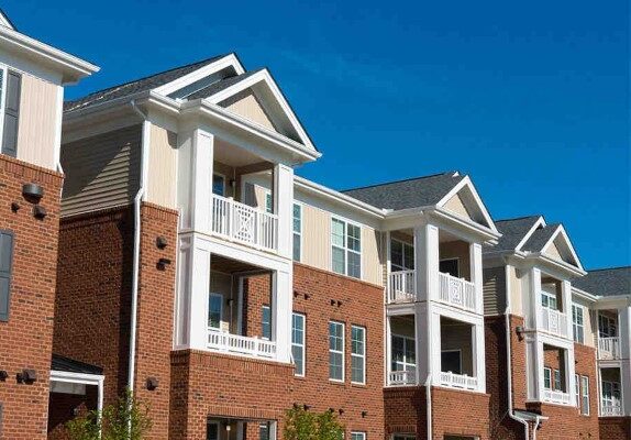 A block of apartments, white on top with balconies and brick below, stand in front of a blue sky.