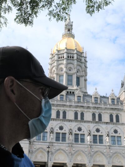 ACLUCT Smart Justice leader Sean Sellars is in the foreground, wearing a mask and baseball hat, looking toward the CT capitol building. In the background is the Connecticut capitol building and a blue sky.