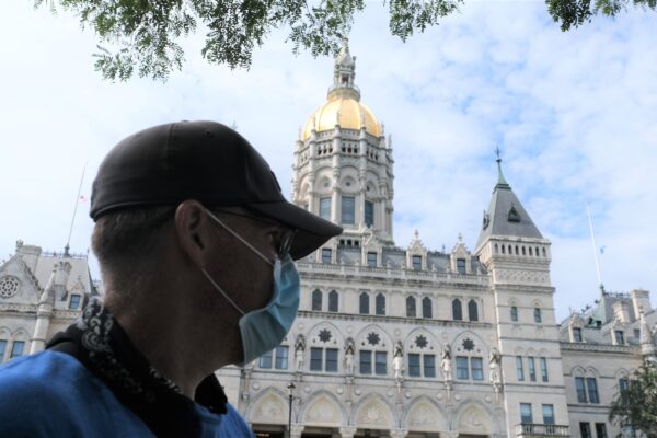 ACLUCT Smart Justice leader Sean Sellars is in the foreground, wearing a mask and baseball hat, looking toward the CT capitol building. In the background is the Connecticut capitol building and a blue sky.