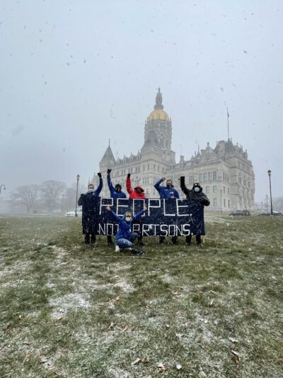 A photo of the Connecticut State Capitol and ACLU-CT Smart Justice leaders with a blue "people not prisons banner." Foreground: capitol lawn, dusted w snow. Middleground: 6 leaders, fists up, hold the banner. Background: snowy sky and capitol building