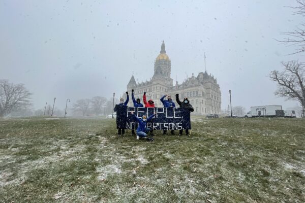 A photo of the Connecticut State Capitol and ACLU-CT Smart Justice leaders with a blue "people not prisons banner." Foreground: capitol lawn, dusted w snow. Middleground: 6 leaders, fists up, hold the banner. Background: snowy sky and capitol building
