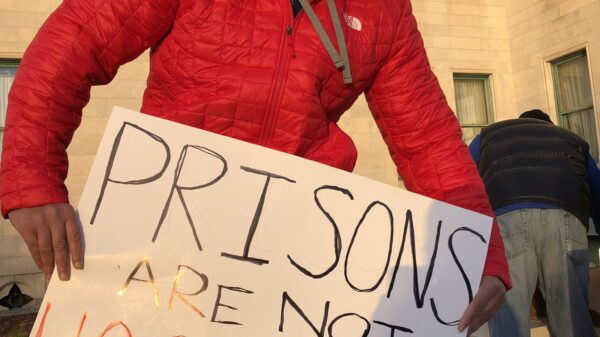 A person in a red coat, black facemask, and black hood holds a sign that says "Prisons are not hospitals" in front of the Connecticut State Capitol building in December 2020.