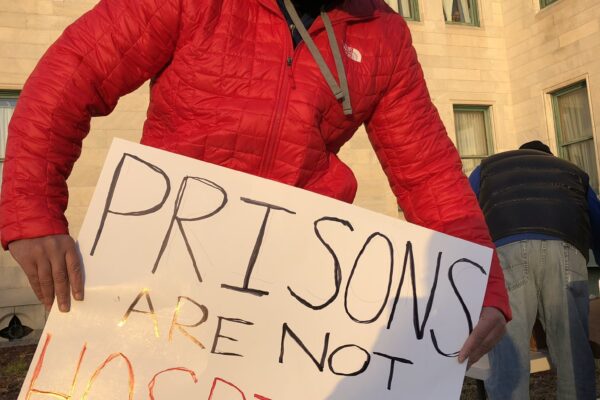 A person in a red coat, black facemask, and black hood holds a sign that says "Prisons are not hospitals" in front of the Connecticut State Capitol building in December 2020.