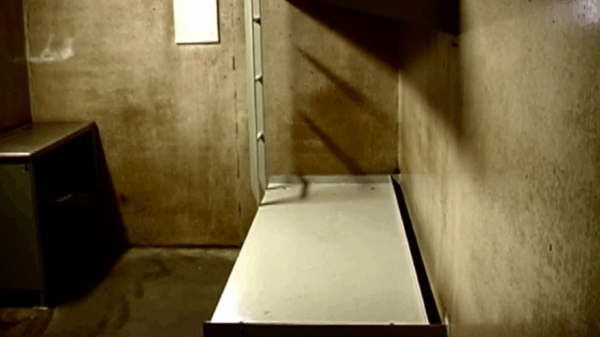 A photo of a bed and desk in a cell at Northern Correctional Institution in Somers, Connecticut. A metal cot-like bed is against a concrete wall on the right. On the left, a small desk and door. The space is small, barren, concrete.