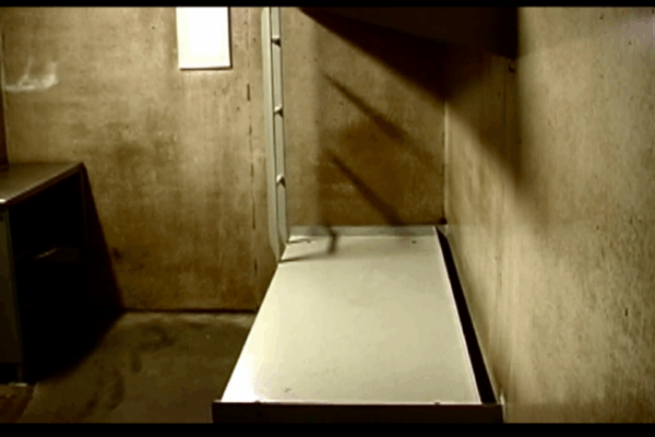 A photo of a bed and desk in a cell at Northern Correctional Institution in Somers, Connecticut. A metal cot-like bed is against a concrete wall on the right. On the left, a small desk and door. The space is small, barren, concrete.