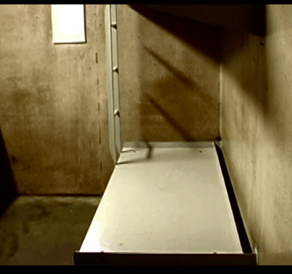 A photo of a bed and desk in a cell at Northern Correctional Institution in Somers, Connecticut. A metal cot-like bed is against a concrete wall on the right. On the left, a small desk and door. The space is small, barren, concrete.