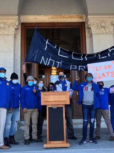 ACLU-CT Smart Justice leaders stand with "people not prisons" banner and "Clean Slate" sign outside of the CT State Capitol. They are behind a podium, and wearing blue "people not prisons" shirts. Many have fists up in power salutes.