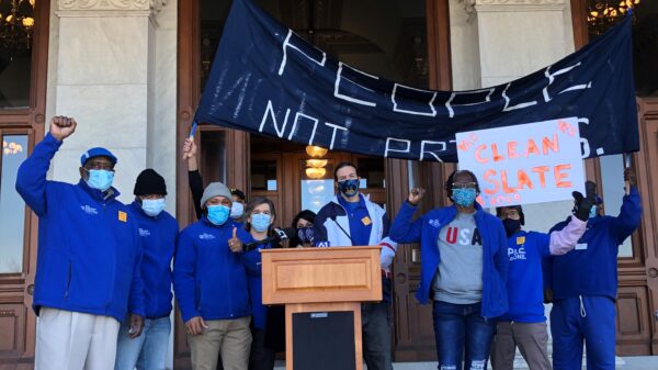 ACLU-CT Smart Justice leaders stand with "people not prisons" banner and "Clean Slate" sign outside of the CT State Capitol. They are behind a podium, and wearing blue "people not prisons" shirts. Many have fists up in power salutes.