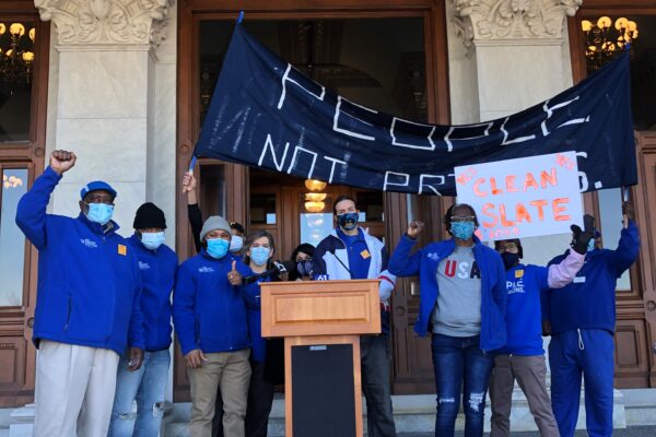 ACLU-CT Smart Justice leaders stand with "people not prisons" banner and "Clean Slate" sign outside of the CT State Capitol. They are behind a podium, and wearing blue "people not prisons" shirts. Many have fists up in power salutes.
