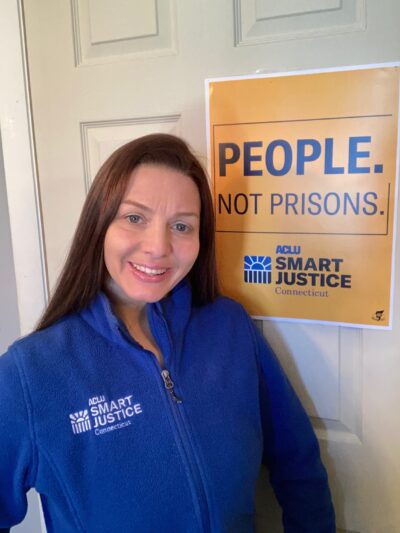 ACLU-CT Smart Justice leader Tracie Bernardi stands in front of a yellow Smart Justice "People Not Prisons" poster. She is wearing a blue ACLU of Connecticut Smart Justice zip up sweatshirt, standing, and smiling.