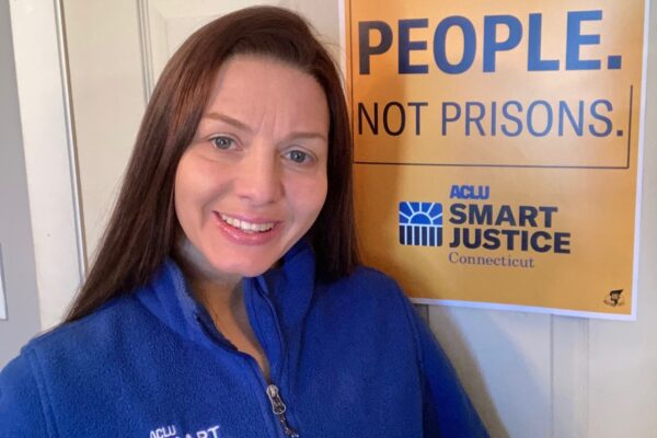 ACLU-CT Smart Justice leader Tracie Bernardi stands in front of a yellow Smart Justice "People Not Prisons" poster. She is wearing a blue ACLU of Connecticut Smart Justice zip up sweatshirt, standing, and smiling.