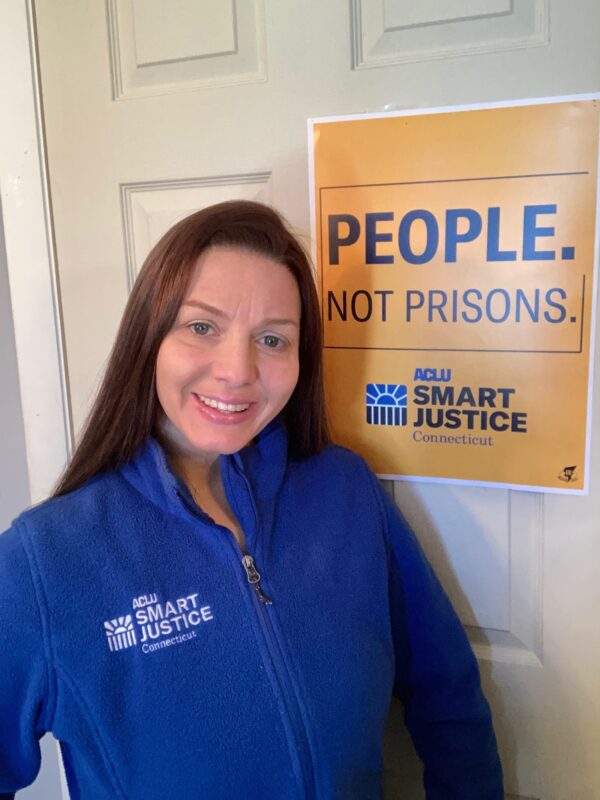 ACLU-CT Smart Justice leader Tracie Bernardi stands in front of a yellow Smart Justice "People Not Prisons" poster. She is wearing a blue ACLU of Connecticut Smart Justice zip up sweatshirt, standing, and smiling.