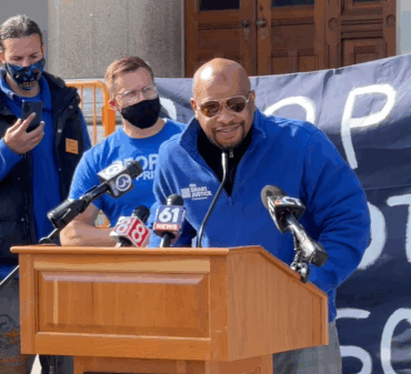 Smart Justice leader Donald Rivers, in a blue zip up Smart Justice shirt and sunglasses, speaks at a podium during an April 21, 2021 press conference.