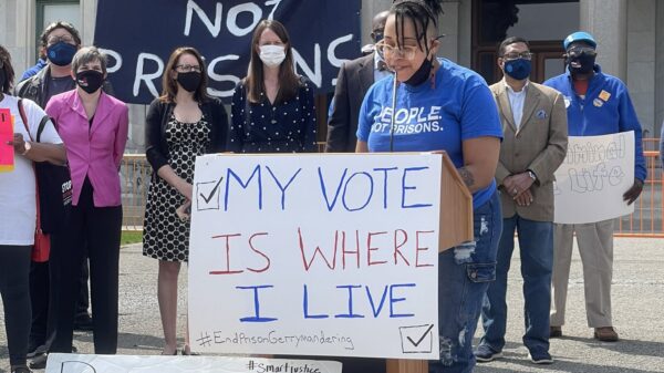 Claudine Fox, ACLU of Connecticut public policy and advocacy director, speaks at a press conference re ending prison gerrymandering. She's at a podium. In front of her is a sign: "my vote is where I live." Behind her, a group of legislators, masked.