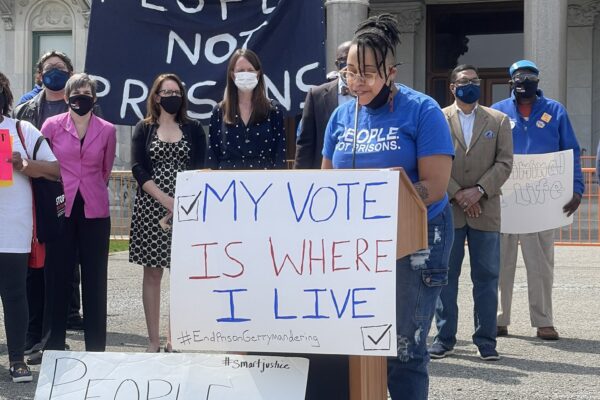 Claudine Fox, ACLU of Connecticut public policy and advocacy director, speaks at a press conference re ending prison gerrymandering. She's at a podium. In front of her is a sign: "my vote is where I live." Behind her, a group of legislators, masked.