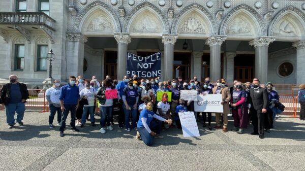 A group of Smart Justice leaders and activists with CONECT kneel and stand in front of the CT Capitol after a Clean Slate press conference. A blue "people not prisons" banner is in the background.