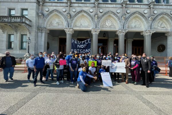 A group of Smart Justice leaders and activists with CONECT kneel and stand in front of the CT Capitol after a Clean Slate press conference. A blue "people not prisons" banner is in the background.
