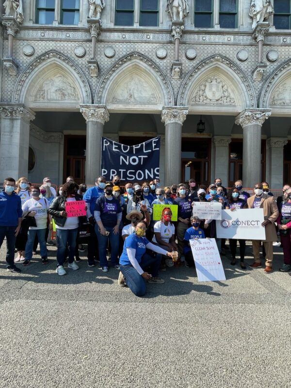 A group of Smart Justice leaders and activists with CONECT kneel and stand in front of the CT Capitol after a Clean Slate press conference. A blue "people not prisons" banner is in the background.