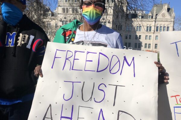 ACLU-CT Smart Justice leader Terri Ricks stands in front of the CT State Capitol Building. She is wearing sunglasses and a rainbow face mask, and she holds a white sign that says, in blue letters, "FREEDOM JUST AHEAD."
