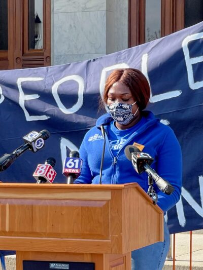 Smart Justice leader shelby Henderson, in a blue Smart Justice zip up and mask, speaks at a podium in front of the CT state capitol. Behind her is a blue people not prisons banner held by two other Smart Justice leaders.