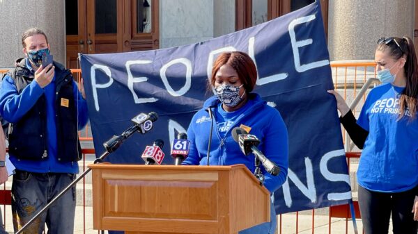 Smart Justice leader shelby Henderson, in a blue Smart Justice zip up and mask, speaks at a podium in front of the CT state capitol. Behind her is a blue people not prisons banner held by two other Smart Justice leaders.