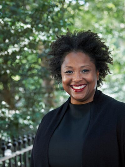 Deborah Archer, national ACLU president, is in the foreground. She is wearing a black shirt and blazer and is smiling, looking directly at the camera. Behind her is a leafy park.