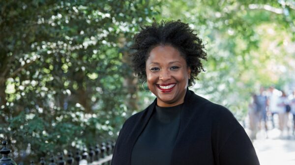 Deborah Archer, national ACLU president, is in the foreground. She is wearing a black shirt and blazer and is smiling, looking directly at the camera. Behind her is a leafy park.