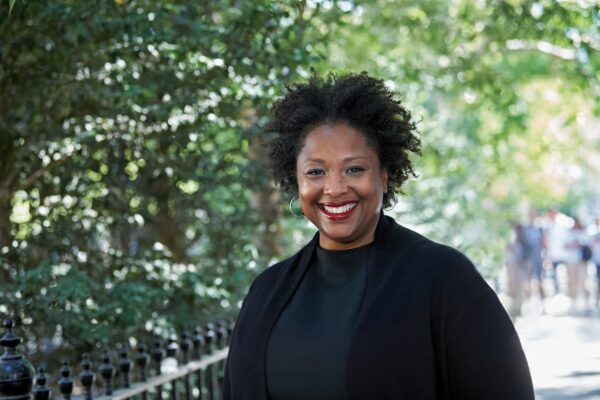 Deborah Archer, national ACLU president, is in the foreground. She is wearing a black shirt and blazer and is smiling, looking directly at the camera. Behind her is a leafy park.