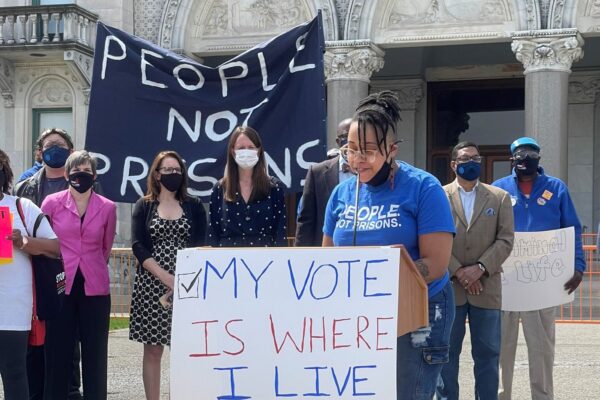 ACLU-CT public policy and advocacy director Claudine Fox, in a blue "people not prisons" smart justice shirt, speaks behind a podium in front of the CT capitol building. The podium sign says, my vote is where I live.