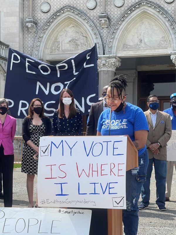 ACLU-CT public policy and advocacy director Claudine Fox, in a blue "people not prisons" smart justice shirt, speaks behind a podium in front of the CT capitol building. The podium sign says, my vote is where I live.