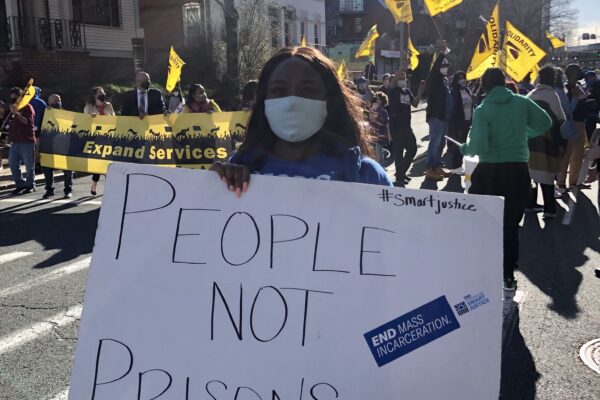 Smart Justice leader Shelby Henderson holds a white sign that says "PEOPLE NOT PRISONS" in blue ink. Behind her is a crowd waving yellow flags and carrying a banner. They are on a street in Hartford, marching.