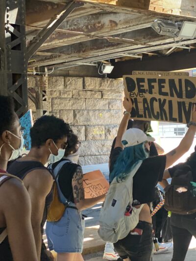 A group of protesters walks in Hartford. One holds a cardboard sign that says, in black letters, "defend Black lives"