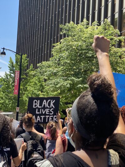 Protesters march in Hartford to call for the state to value Black lives. In the foreground, a person has their fist up. In the background, a Black Lives Matter sign