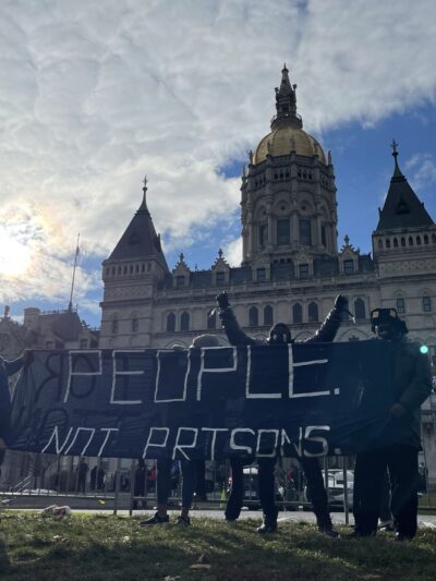 Smart Justice leaders stand in front of the CT capitol. The sun is breaking through a cloud. One person stands with arms upstretched above their head, behind the people not prisons banner