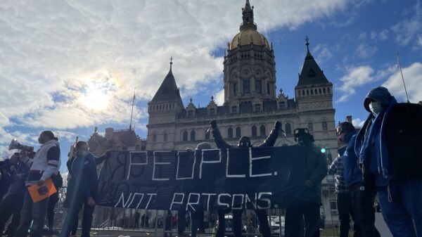 Smart Justice leaders stand in front of the CT capitol. The sun is breaking through a cloud. One person stands with arms upstretched above their head, behind the people not prisons banner