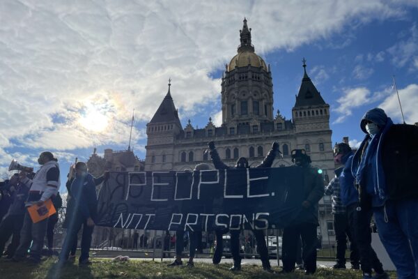 Smart Justice leaders stand in front of the CT capitol. The sun is breaking through a cloud. One person stands with arms upstretched above their head, behind the people not prisons banner