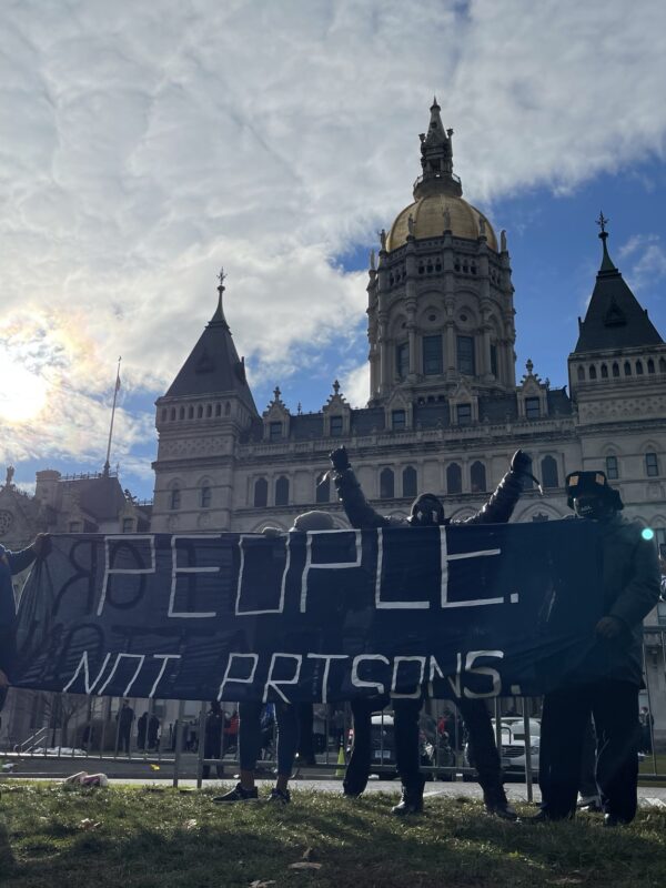Smart Justice leaders stand in front of the CT capitol. The sun is breaking through a cloud. One person stands with arms upstretched above their head, behind the people not prisons banner