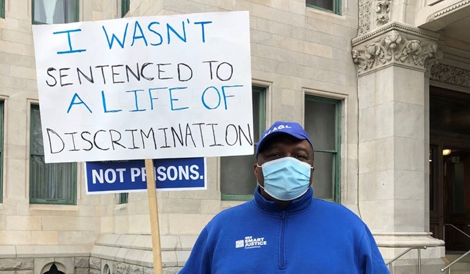 Anderson Curtis, in blue sweatshirt and hat and mask, holds a sign that says